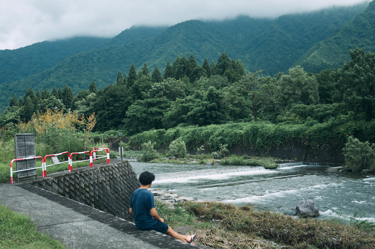 A person sitting beside a river below forested mountains.