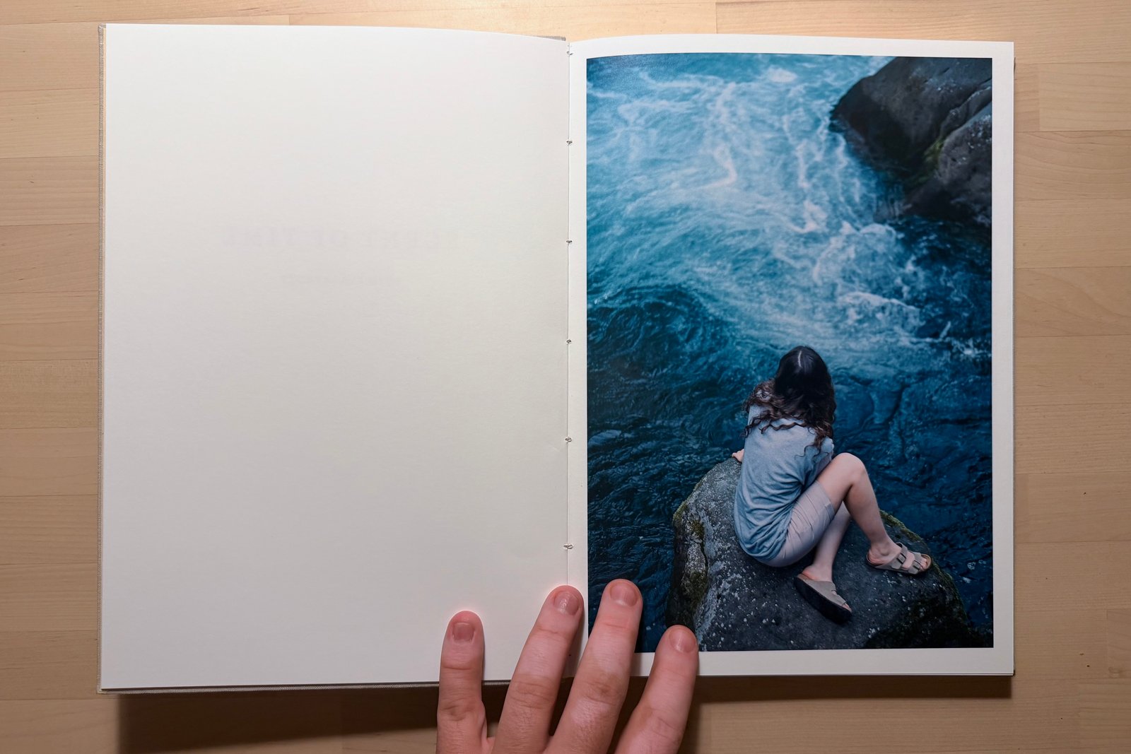The finished Scent of Time maquette open on a wooden table: a blank left page facing a photograph of a person sitting on a rock above rushing blue water.