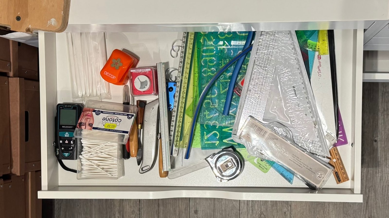 An open drawer in the studio, crowded with rulers, stencils, set squares, a laser distance meter, craft punches, a utility knife, and a tape measure.