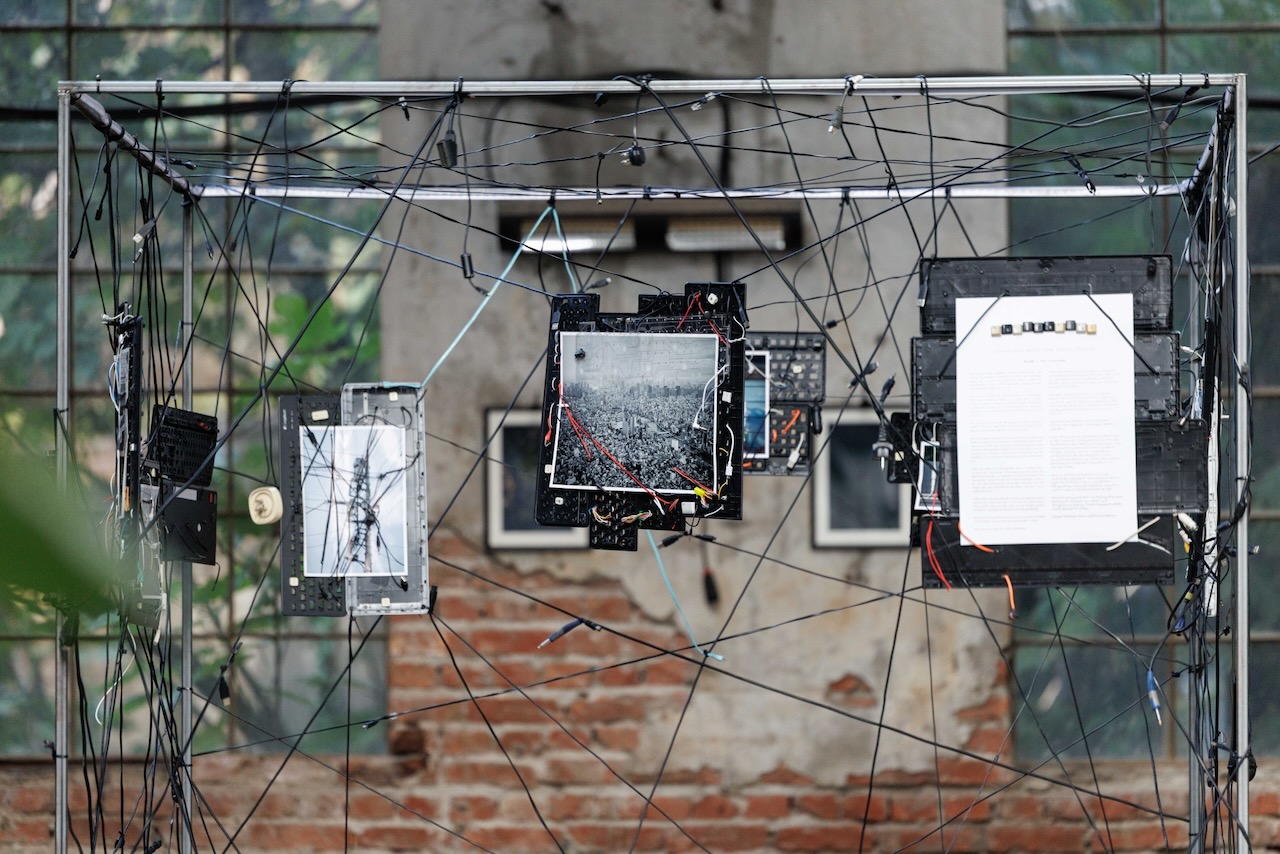 Urban photographs and a wall text held in a web of cables and keyboard parts inside the Wirescapes cube.